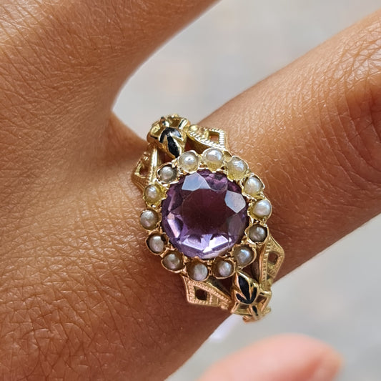 Antiker Gelbgoldring mit verschnörkelter und verzierter Ringschiene welche links und rechts des Ringkopfes mit schwarzer Emaille malerei verziert ist. Das Herzstück des Ringes ist ein fliederfarbener Amethyst umringt von zarten saatperlen 